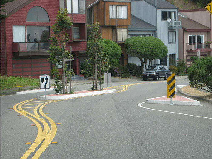 Figure 3.5.5. Chicane in Residential Area. This figure contains a photograph of a residential area. In the background there is a row of houses. In the foreground is a chicane bearing to the right. The center median has plants and right of way signs. A triangular curb extension on the right hand side of the photograph is highlighted by a rectangular sign with yellow and black horizontal lines.