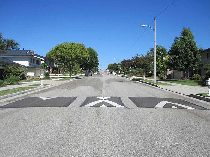 Figure 1.2. Vertical Deflection Measure - Speed Cushion. This figure contains a photograph which illustrates a wide residential street free of traffic. Three speed cushions are present, each a small change in height and painted with white indicator arrows showing right of way.