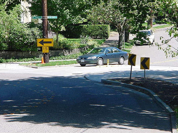 Figure 3.6.3. Corner Extension at Realigned Intersection.  This figure contains a photograph of an intersection which has been realigned using a curb extension. A car in the top right of the picture must curve around the extension to continue past a yellow sign indicating a speed limit of fifteen miles per hour with an arrow pointing to the left. A curb curves from the bottom right of the photograph to the left so that anyone turning right at the intersection must curve around it.