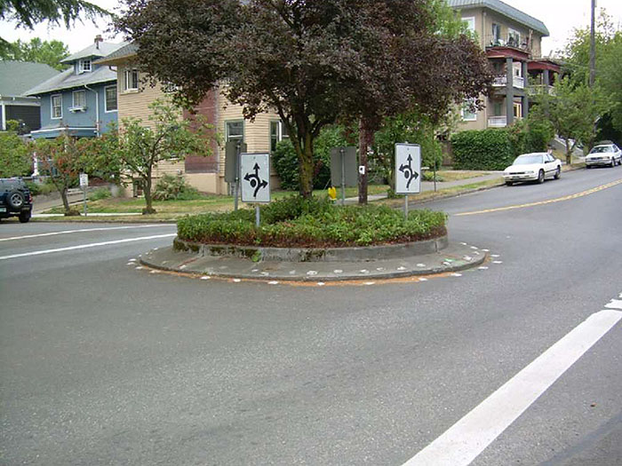 Figure 3.7.1. Landscaped Traffic Circle. This figure contains a photograph of a traffic circle in a residential neighborhood. The concrete circle has white dots following its circumference. Low plants and a tree fill its center. White, square signs facing each section of street show that traffic should keep right and the points at which the circle can be exited using curved lines and arrows.