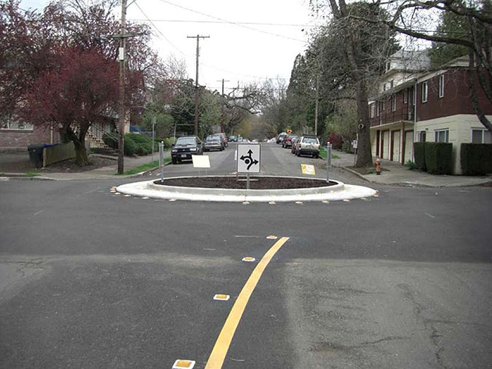 Figure 3.7.2. Traffic Circle without Landscaping. This figure contains a photograph of a traffic circle in a residential neighborhood. The concrete circle has white dots following its circumference. The center is clear with no landscaping and allows a clear line of sight. White, square signs facing each section of street show that traffic should keep right and the points at which the circle can be exited using curved lines and arrows.