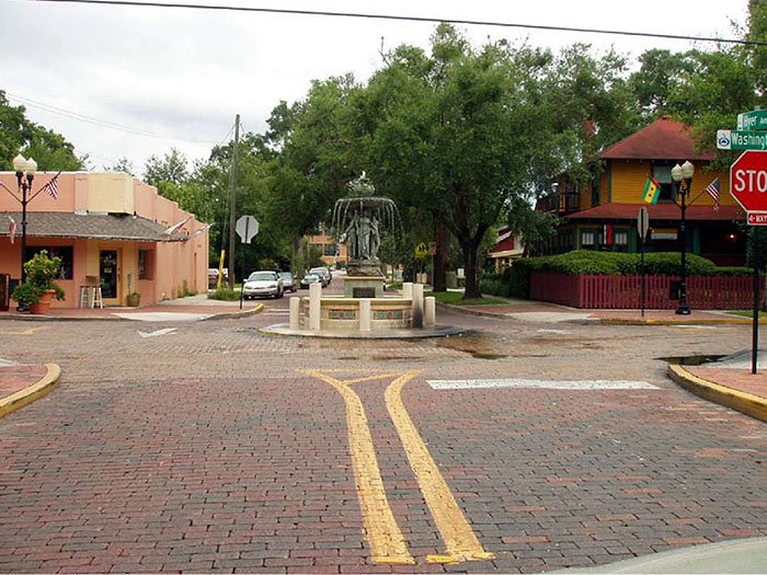 Figure 3.7.3. Traffic Circle in Commercial Setting. This figure contains a photograph of a traffic circle in a commercial setting. The streets are brick and there is a stop sign at each corner. The circle has concrete pillars around its circumference. A fountain fills its center. There are trees in the picture's background and businesses on each far corner of the street.