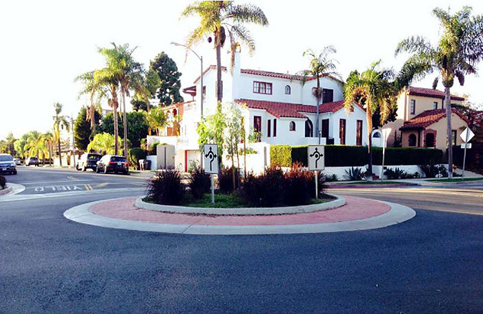 Figure 3.7.4. Traffic Circle In Residential Setting. This figure contains a photograph of a traffic circle in a residential neighborhood. The concrete circle has a red brick walk following its circumference. The center is well landscaped with grass and low, red bushes. White, square signs facing each section of street show that traffic should keep right and the points at which the circle can be exited using curved lines and arrows. On the corner opposite there is a white stucco house and palm trees.