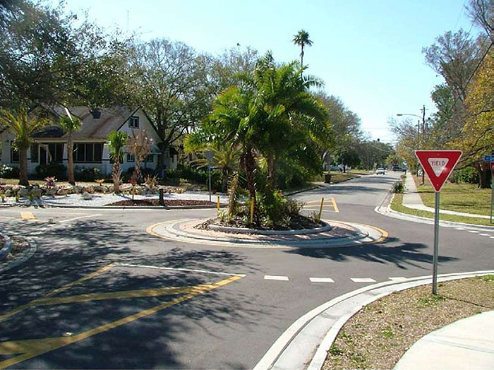 Figure 3.8.1. Small Modern Roundabout. This figure contains a photograph of a traffic circle in a residential neighborhood. The concrete circle has red brick around its circumference. The center is landscaped with palm trees. The street in the bottom left hand corner of the picture curves to the right. Dotted lines and yield signs are at each section of street. A house can be seen in the upper left hand corner of the picture.