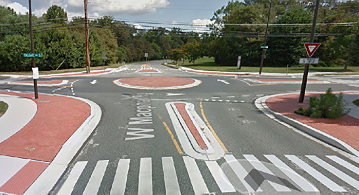 Figure 3.8.2. Mini-Roundabout with Splitter Islands. This figure contains a picture of a small red brick roundabout. Each section of road leading off from it has small red brick islands to split the road into lanes. The corners have yield and street signs.