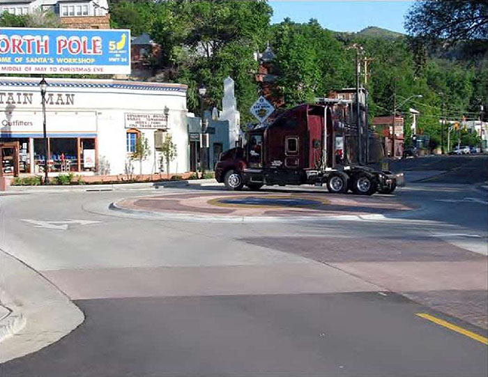 Figure 3.8.3. Mini-Roundabout Center Island with Color Pavement. This figure contains a picture of a mini-roundabout with pink and brown pavement. A semi-truck is traversing the roundabout near a store.