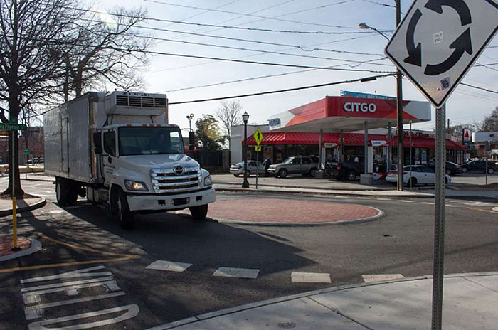 Figure 3.8.4. Mini-Roundabout with Truck. This figure contains a picture of a mini-roundabout, colored red. A freezer truck is traversing the roundabout. A white sign is in the foreground, depicting a circle made of arrows on a white background. A gas station is in the background.