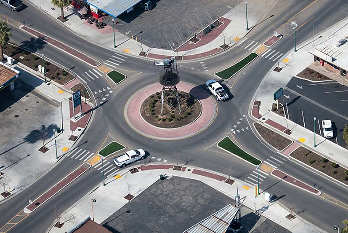 Figure 3.9.1. Single-Lane Roundabout. The figure contains an overhead photograph of a single lane roundabout. The splitter islands are green and red. The roundabout contains a tower surrounded by low bushes. Two pickup trucks are using the roundabout; one waiting to enter and the other in circle.
