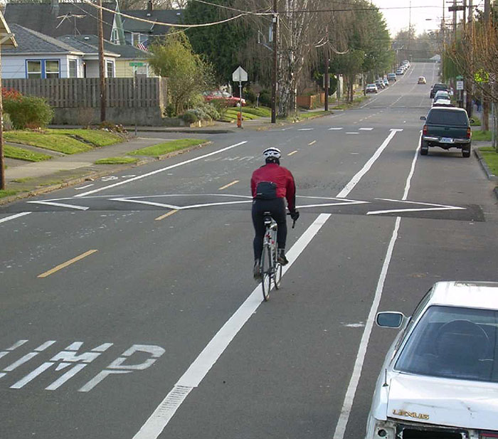 Figure 3.10.1. Speed Hump with Bicycle Lane and On-Street Parking. The figure contains a photograph of a two lane road with a bicycle lane and on-street parking. A bicyclist in the bicycle lane is moving away from the photographer and approaches the speed hump, a raised portion of pavement painted with white angular lines. Cars and trucks are parked in a parking lane on the right hand side of the road.