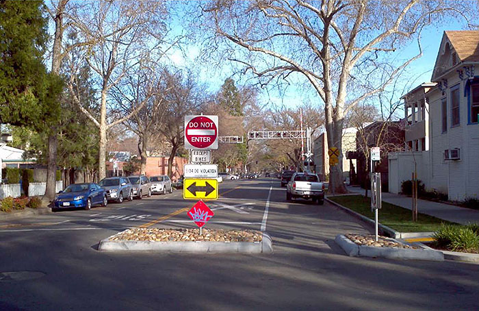 Figure 1.4. Routing Restriction Measure - Half Closure. This figure contains a photograph which illustrates a half closure, restricting access down a two lane residential street to bicycles only. A concrete square filled with rocks is a physical barrier. A signpost holds four street signs (from top to bottom): a square white sign with a red circle containing the words Do Not Enter, a white rectangular sign with the words Except Bikes, a yellow sign with a double headed black arrow pointing left to right, and a red triangle covered in blue graffitti. There is also a railroad crossing in the near distance.