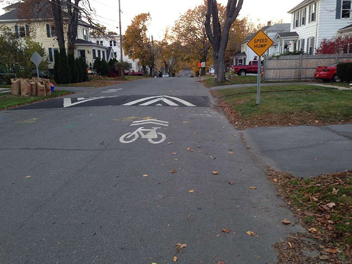 Figure 3.10.2. Speed Hump on Residential Neighborhood Street. This figure contains a photograph of a street in a residential area. A speed hump is marked by a yellow diamond sign and painted white angular lines. A painted bicycle icon and arrows are painted on the street just before the hump.
