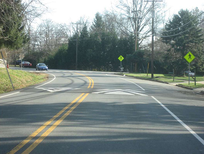 Figure 3.10.3. Speed Hump Along Horizontal Curve. This figure contains a photograph of a curving two lane road divided by a double yellow line in a wooded area. Just before the curve a speed hump is in place a short distance from a cross walk. The crosswalk is indicated by green diamond signs on the right hand side of the road.