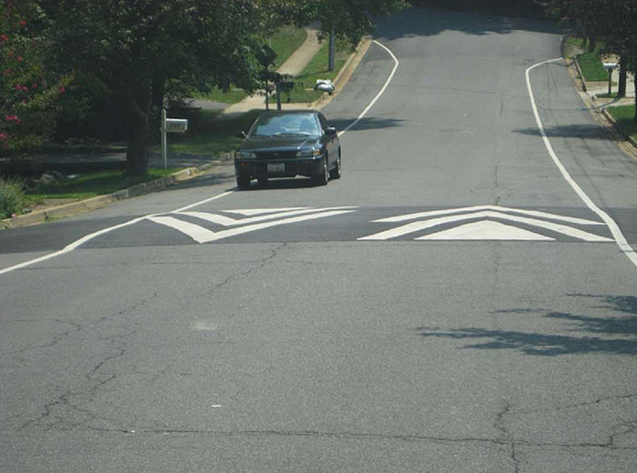 Figure 3.10.4. Speed Hump on a Grade. This figure contains a photograph looking down a hill. The two lane, unmarked road, has an oncoming car in the left lane just about to cross over a speed hump.