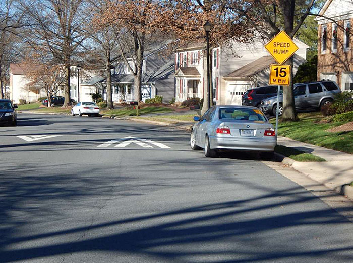 Figure 3.10.6. On-Street Parking Adjacent to Speed Hump. This figure contains a photograph of a curved street in a residential area. A car is parked near the curb beside a signpost with a yellow diamond speed hump sign and a 15 MPH speed limit sign. Other cars can be seen parked on either side of the street.