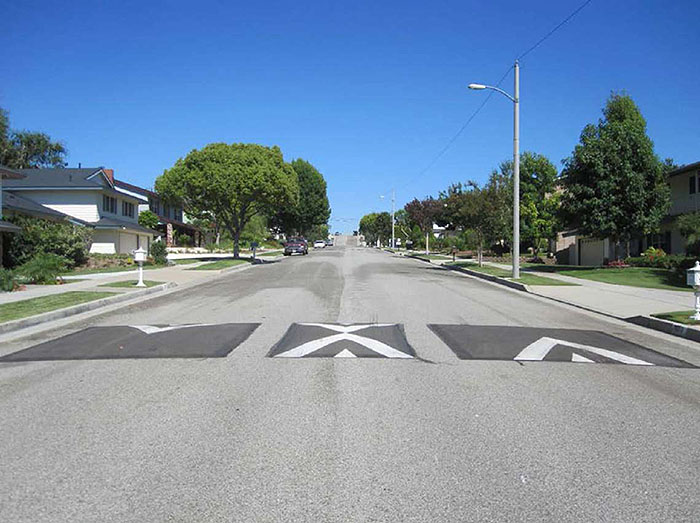 Figure 3.11.1. Speed Cushion with Passage that Straddles Centerline. This figure contains a photograph of a tree lined undivided street in a residential area. Three speed cushions are visible, crossing the road from right to left. The right and left cushion are painted with wide white angular stripes indicating a speed cushion. The cushion which straddles the centerline has angular stripes on each side and is narrower than the other two.