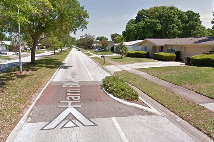 Figure 3.12.2. Speed Table with Choker. This figure contains a photograph of a residential area. On the left hand side of the photo there is a lane of trees on a landscaped median. To the right of the trees there is single lane road labeled Harn Blvd. A speed table, dyed a dusty pink, fills most of the lane. This traffic feature also uses a choker, a crescent shaped curb extension landscaped with shrubs that extends onto the speed table. To the right of that, there is a sidewalk separating the street from a row of single story houses.