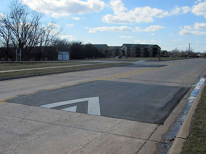 Figure 3.13.1. Offset Speed Table. This figure contains a photograph of a straight two lane road divided by a double yellow line. In the near distance in the right lane only, there is a speed table with striping indicating the right of way. A car length further away, there is an offset speed table in the left hand lane. Raised reflectors separate the lanes in between the two speed tables. Off of the left hand side of the street there are apartments in the distance.