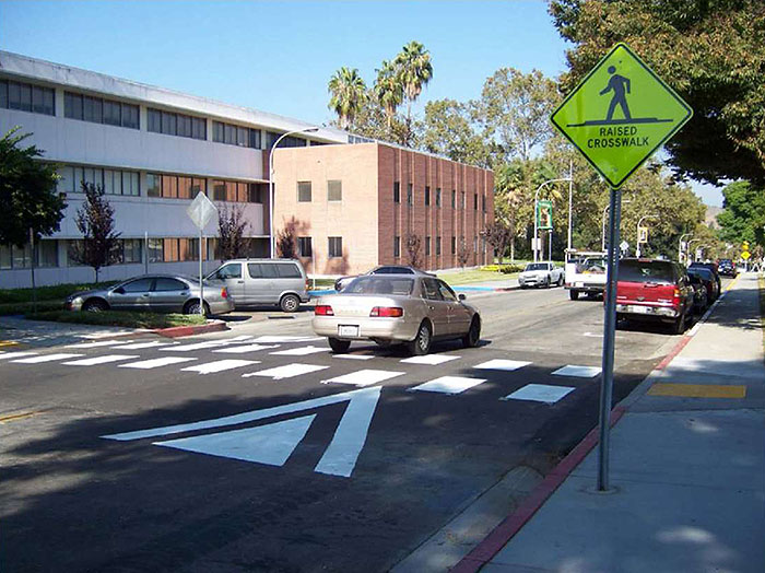 Figure 3.14.2. Typical Raised Crosswalk Application. This figure contains a photograph of a two lane street in a business district. A sidewalk on the right hand side of the picture has a signpost holding a diamond shaped yellow sign showing an icon of a person walking over the words Raised Crosswalk. Between the curb and the right lane there is parallel parking where a red truck and another vehicle are parked. A light colored car and another truck have just crossed the walk in the right hand lane. The left lane is free of traffic. A raised crosswalk connects the sidewalks on the sides of the street.