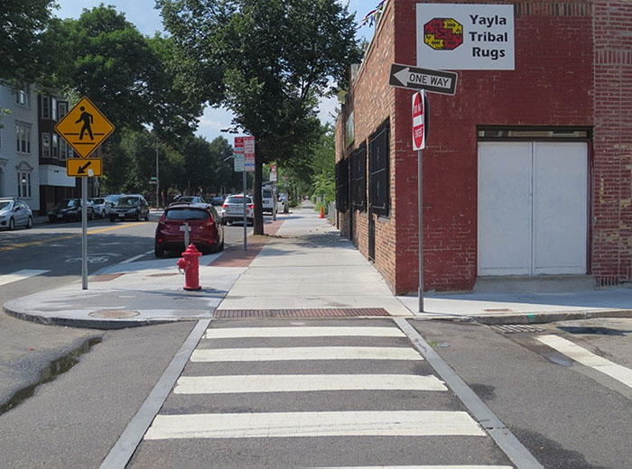 Figure 3.14.4. Raised Crosswalk at Intersection. This figure contains a photograph of a raised crosswalk at an intersection taken from the point of view of a pedestrian. The white striped pattern crosses to a sidewalk which stretches up the center of the photo. To the right of the sidewalk, there is a red brick single story building. A signpost to the right of the crosswalk just at the curb holds a Do Not Enter sign and a One Way sign pointing to the left. Across the sidewalk from that there is a red fire hydrant and a crosswalk sign. Trees and business line the street to the left of the sidewalk.