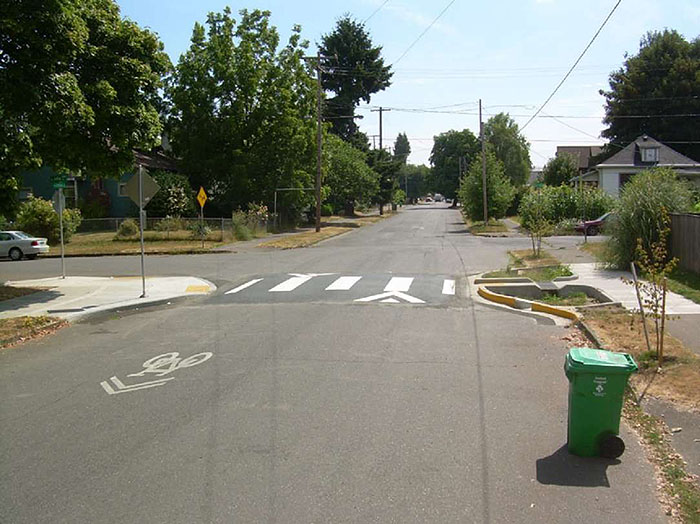 Figure 3.14.5. Raised Crosswalk with Curb Extension. This figure contains a photograph of a tree-lined residential area. The focus is a road which runs from the bottom middle of the photo up to the top middle. The road narrows at approximately the halfway point using a curb extension. There is a raised crosswalk that leads from the curb extension to the opposite corner of a sidewalk. Just above the crosswalk, there is an intersection.