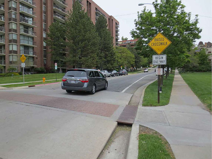 Figure 3.14.7. Raised Crosswalk Design to Accommodate Drainage. This figure contains a photograph of a street from the bottom left to the top right of the photo. The raised crosswalk has pale concrete ramps and a red brick crosswalk. A gutter allows drainage on the right hand side of the crosswalk and a metal plate covers it to allow foot traffic to easily use the crosswalk. A signpost between the crosswalk and sidewalk holds a yellow diamond sign which says Raised Crosswalk and a white rectangular sign which says Begin One Way. A sidewalk runs beside the road separated from it by a row of trees. A van is crossing the walk and across the street there is a large red brick apartment building. Space for parallel parking is available on either side of the street