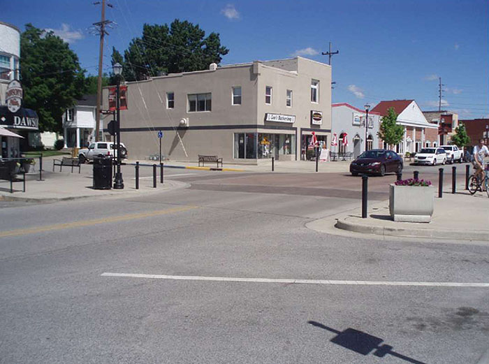 Figure 3.15.1. Raised Intersection. This figure contains a photograph of a commercial area including a two lane street running from bottom left to upper right of the photo. The raised intersection, a speed table covering the entire intersection, dominates the center of the photo. The ramps are darker and the plateau is a light red. To the right of the raised intersection there is a sidewalk and a concrete flower box filled with a dark red and green plant. To the left of the intersection there are a row of shops. One car is moving through the intersection and there are cars parked in parallel behind it.