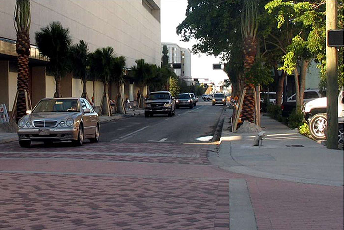 Figure 3.15.2. Raised Intersection in Dense Urban Setting. This figure contains a picture of a two lane, one way road between two strands of palm trees and multi-story buildings. There is oncoming traffic in both lanes. Two entrances to the raised intersection can be seen. The intersection itself is done in light red brick.