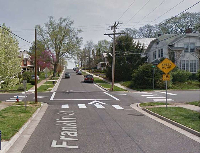 Figure 3.15.3. Raised Intersection in Residential Setting without Raised Crosswalks. This figure contains a photograph of undivided two lane roads intersecting at the center of the photo. The road running from top to bottom, Franklin St., features parallel parking on the right hand side and a car approaches the intersection. There is a signpost to the right of Franklin, at the intersection, with a yellow diamond sign with the words Speed Hump and a square yellow sign below that which contains the text 15 MPH. There are houses on the opposite side of the intersection. Arrows painted between the crosswalks and the raised pavement indicate right of way. The raised intersection is bordered in white stripes and sidewalks.