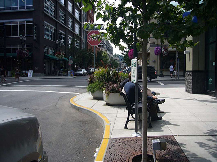 Figure 3.16.3. Corner Extension in Urban Setting. This figure contains a photograph with a focus on the corner extension. Near the bottom of the picture there is a tree planted at the curb. Further up the sidewalk on the corner extension there is a bench with people seated on it and a series of large, round planters. There is a stop sign at the intersection facing the camera. A four story building occupies the corner diagonally from the extension. The opposite corner has a set of shops.