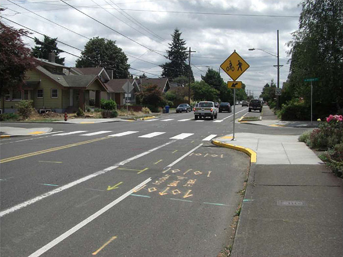 Figure 3.16.5. Corner Extension and Bicycle Lane. This figure contains a photograph of a street running from the bottom left to the top right. Another street forms an intersection running across the photo's centerline. A signpost can be seen on the corner extension extending into the street running top to bottom. A diamond shaped yellow sign depicts an icon of a person walking and a bicycle. Near the curb there is space for parallel parking and between the parking and the lane there is a separate bicycle lane. Crosswalks are visible running left to right at the intersection. A row of houses can be seen on the corner opposite.