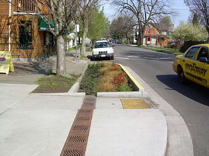 Figure 3.16.6. Corner Extension with Bioswale. This figure contains a photograph focusing on a corner extension. The center of the extension contains a rectangular depression filled with grass and low bushes. To the left of this planting box there is a tree growing from another dedicated planting area. The section of the extension near the bottom of the photo has a ramp cut to street level. A white vehicle is parked parallel to the curb on the far side of the extension. A yellow taxi can be seen driving off camera towards the right side of the picture. A series of shops can be seen on the left hand side of the picture.