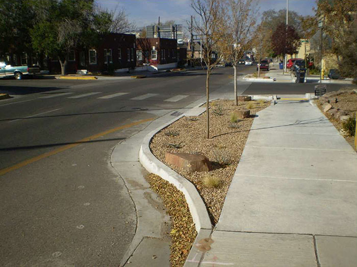 Figure 3.16.8. Corner Extension with Drainage Structure Relocation. This figure contains a photograph of a street running from the bottom left to the center of the top edge. A sidewalk and corner extension dominate the bottom right hand quadrant of the picture. The corner extension is filled with rocks and small trees. Dark, one story buildings occupy the opposite corner of the intersection. This corner extension has a drainage structure in it.