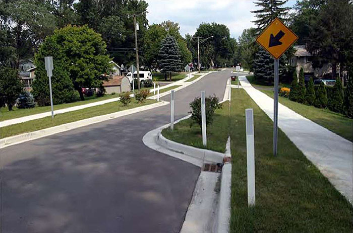 Figure 3.17.2. Angled Choker. This figure contains a photograph of a street running top to bottom near the center of the picture with an angled choker. A rounded curb extension on each side of the road causes it to narrow and angle from left to right. The extensions are covered in grass and are separated from nearby houses by sidewalk. White reflector posts follow the curved portion of each extension. A signpost on the right side of the street contains a diamond shaped yellow sign with a black arrow pointing down and to the left.