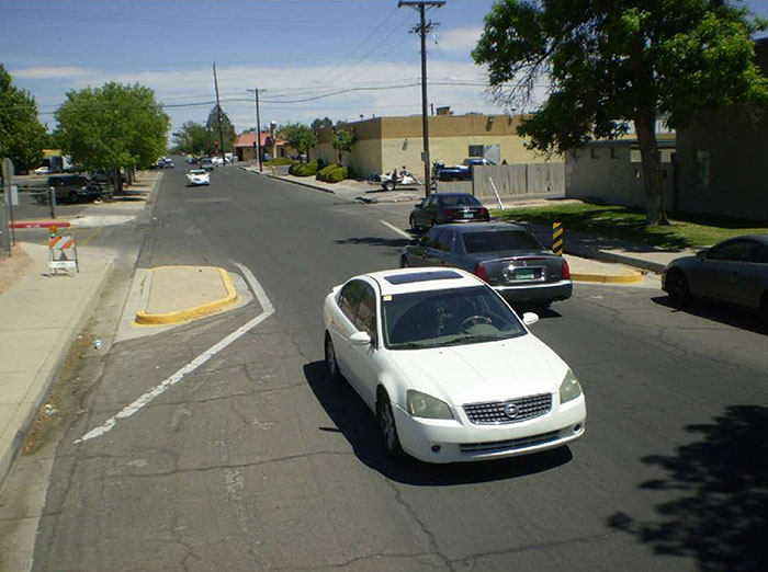 Figure 3.17.5. Choker with Passing Traffic. his figure contains a photograph of an undivided two lane street running from the bottom right to near the top left of the picture. Curb extensions, detached from the sidewalks and outlined in white stripes, dominate the center of the photograph. A white car travelling towards the camera and a gray car travelling away are passing through the choker at the same time.  A car is parallel parked in the opposite lane, pointed away from the camera after the chocker. Trees are scattered around the sides of the street and a  tan building can be seen in the distance.