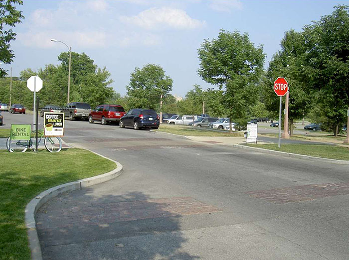 Figure 3.17.6. Choker with Pedestrian Crosswalk. This figure contains a photograph of a two lane street running from bottom right to top left. A choker is used to narrow the street in the center of the photo for a crosswalk. The extensions are landscaped with grass. A stop sign can be seen on the right hand extension, near a sidewalk. Cars are parallel parked on the right hand side of the street. On the far side of those cars there is a parking lot. On the left hand side of the street, on the extension near the intersection, there is a small green sign for bike rentals.