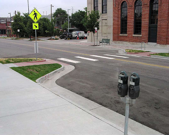 Figure 3.17.7. Choker Adjacent to On-Street Parking. This figure contains a photograph of a two lane street divided by a double yellow line, using a choker at a crosswalk. The left hand side of the street shows available parallel parking near two parking meters. Buildings and off street parking can be seen across the street. The curb extensions are landscaped with grass on either side of the ramps to the sidewalk.