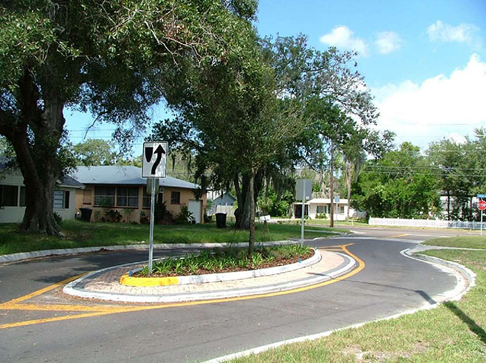 Figure 3.18.3. Landscaped Oval Median Island. This figure contains a photograph of a street running through a residential area. The only part of the street that's visible is the section using an oval median island to narrow the lanes. The island is landscaped with a tree and low ground cover. A white sign indicates right of way around the island. Several houses are visible across the street from the camera.