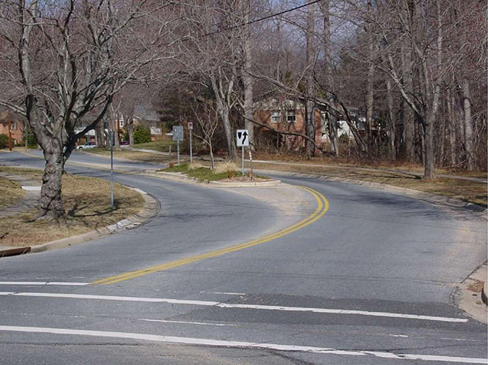 Figure 3.18.4. Median Island Near Curve at Entrance to Residential Area. This figure contains a photograph showing the entrance to a residential area. As the road curves to the left, a median island is used to narrow the lanes. The island contains some landscaping and street signs. The only clearly visible sign is the one indicating right of way. Bare trees line either side of the street. Two houses are visible through trees in the distance.