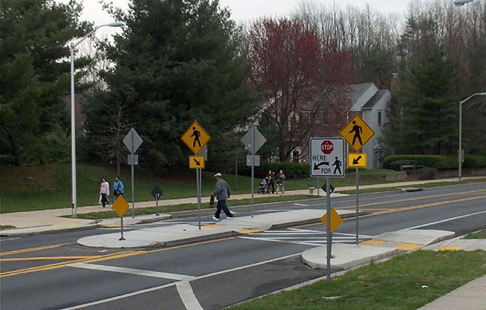 Figure 3.18.5. Median Island with Crosswalk. This figure contains a photograph of a divided two lane street. Focus is on an island with a crosswalk. Curb extensions are also used to narrow the lanes near this island. A pedestrian is crossing from right to left and is just leaving the island to cross the far lane. Two other pedestrians are walking on the far sidewalk. A white, square sign on the near side of the street shows that cars should stop at the crosswalk for pedestrians. A yellow diamond shaped sign with the icon of a pedestrian and a square sign with an arrow points to the crosswalk. Similar signs occupy the island and far sidewalk.