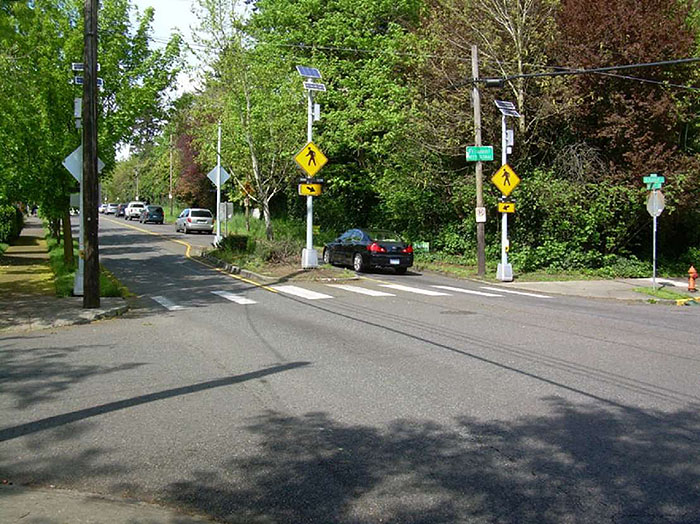 Figure 3.18.6. Median Island at Intersection. This figure contains a photograph of a median island at an intersection. The road leading from right to left occupies the bottom half of the picture. A crosswalk runs parallel to to this road. The street intersecting it uses a landscaped median to narrow the lanes. Two pedestrian crossing signs can be scene near the island. A car is driving away in the right lane. Additional traffic can be seen in the distance in the right lane. Trees and undergrowth can be seen on either side of the street.