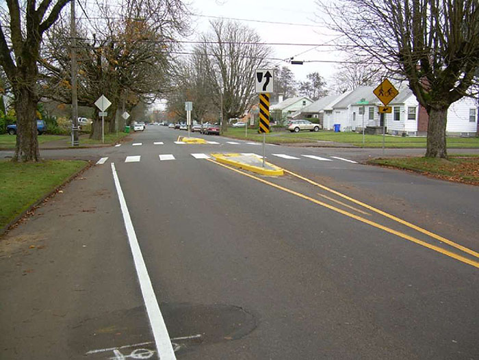 Figure 3.18.7. Small Median Islands at Intersection. This figure contains a photograph focusing on a road running from bottom to top. An intersection in the top third of the picture shows the use of small median islands before and after the intersection to narrow the lanes. The islands are rounded triangles of concrete containing yellow and black-striped  signs and white signs showing right of way. Large trees line either side of the streets and houses can be seen on the far right side of the street.