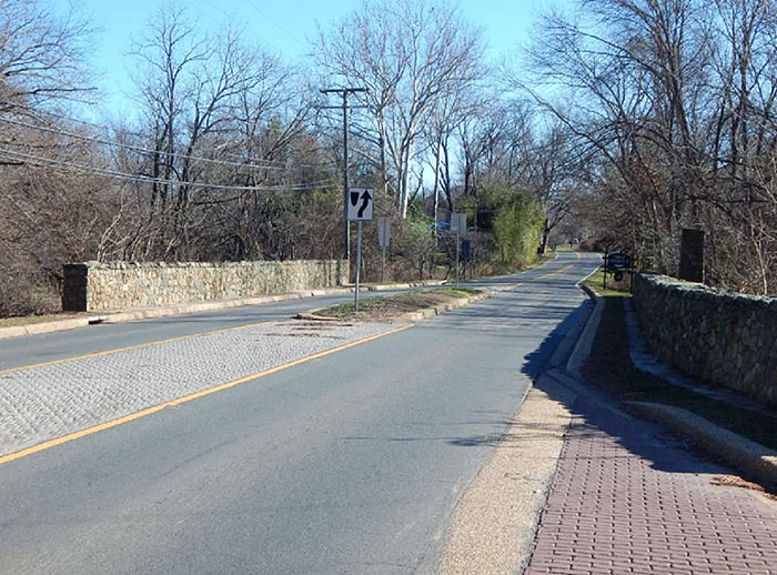 Figure 3.18.8. Short Midblock Median Island. This figure contains a photograph of a two lane street which uses a median island to narrow the lanes where there are sections of stone walls on either side of it. Trees and heavy undergrowth fill either side of the street beyond the walls.