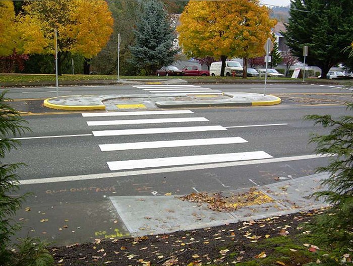 Figure 3.18.10. Median Island with Pedestrian Refuge and Offset Crosswalk. This figure contains a photograph of a four lane road running from left to right. The section of crosswalk traversing the nearest two lanes leads to a large, oval island with a recessed sidewalk. The walkway curves to the right along the island's length and the crosswalk resumes, offset to the right. A small parking area can be seen across the street along with a number of trees.