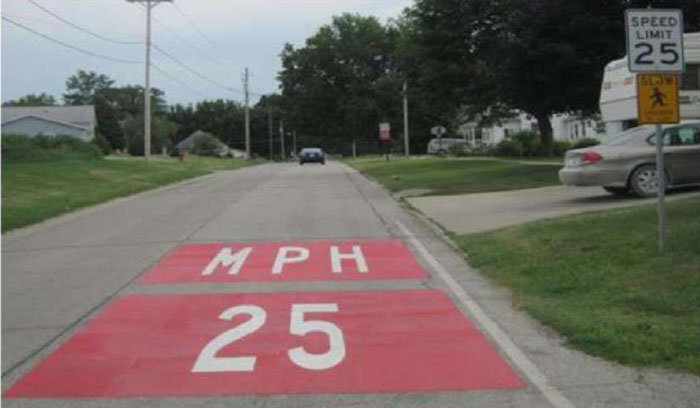 Figure 6-2 Horizontal signing at community entrance in Ossian, IA [Treatment authorized with MUTCD experimental waiver] (Image Source: Hallmark, et al 2002). This photo shows an experimental treatment with 25 MPH painted in red on the road. Note: The red background coloring is not MUTCD compliant, and should only be installed after receiving approval from FHWA through an experimentation waiver.