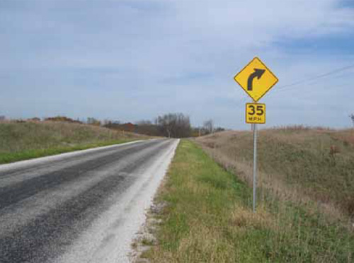 Photo. A rural roadway is shown with a horizontal curve warning sign on the shoulder. The sign consists of two separate components; the top is a yellow diamond with a black curve arrow and the second is a smaller square plaque displaying 35 m.p.h.