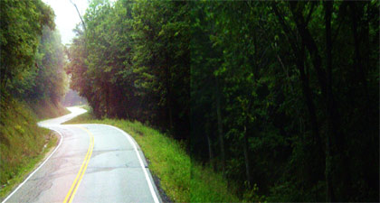 Photograph - Photograph displaying a rural two-lane highway with no useable shoulder and no useable clear zone, considering a steep slope, trees, and roadside rock cliff.