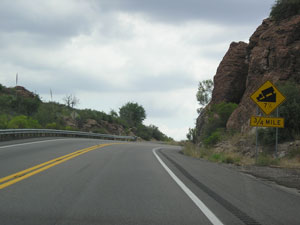 The photo shows a roadway going up a mountain area where it curves around to the right. There is a sign on the right alerting trucks to an upcoming steep grade down.