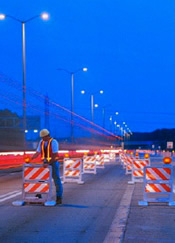 worker positioning a barricade: he is wearing a hardhat and vest with horizontal and vertical reflective stripes